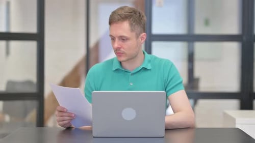 Young Adult Reviews Documents at Desk with Laptop