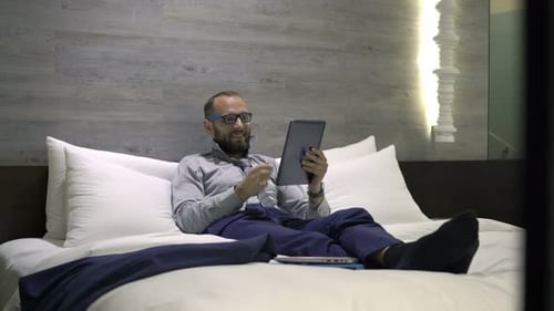 Businessman Chatting On Tablet Lying On Bed In Hotel Room At Night