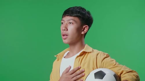 Side View Of Asian Boy With A Ball Singing National Anthem While Cheering Soccer On Green Screen