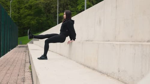 A Beautiful Darkhaired Sporty Girl is Doing Leg Exercises Outdoors in the Park on the Stairs