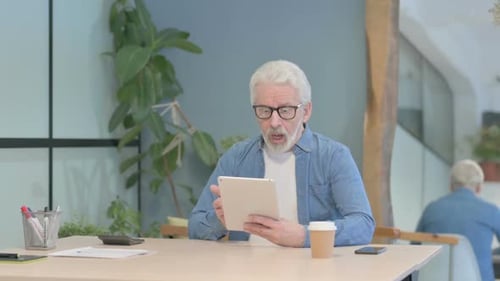 Gray Haired Man Looks at Tablet at Desk