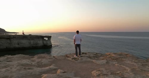 Aerial View of a Man Standing on the Edge of a Cliff Near the Sea at Sunset