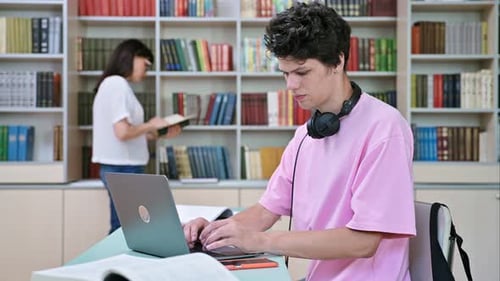 Jeune étudiant de sexe masculin assis avec un ordinateur portable avec des livres dans la bibliothèque du collège