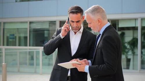 Two Businessmen Standing Outside a Modern Office Building Using a Tablet for Discussing Work
