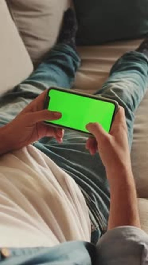 Young man lying on sofa uses mobile phone with green mock-up screen chroma key