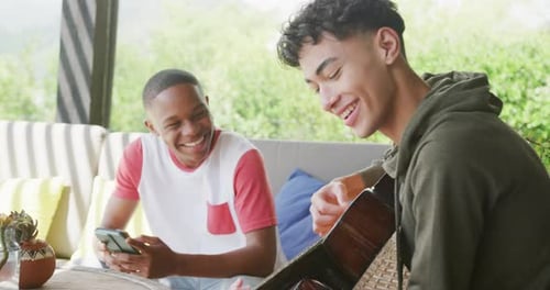 Happy diverse male teenage friends playing guitar at home, slow motion