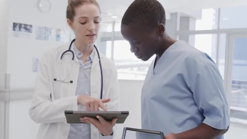Diverse female doctor and nurse looking at tablets and talking in hospital corridor, in slow motion