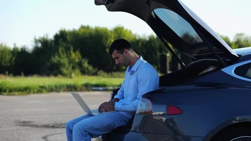 Young Handsome Entrepreneur Works on Notebook Sitting in Electric Car Trunk Successful Businessman