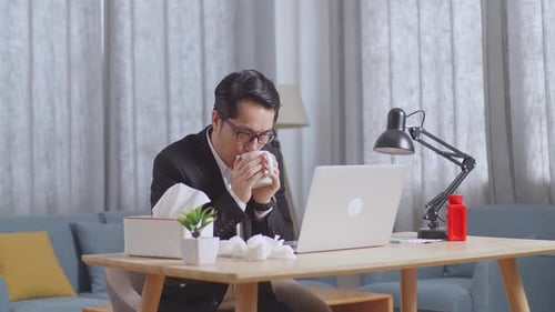 Sick Asian Businessman Drinking Hot Water While Working With Laptop In The Living Room At Home