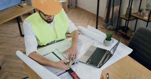Top view. Man designer in helmet and vest which working with architectural scheme on blueprint