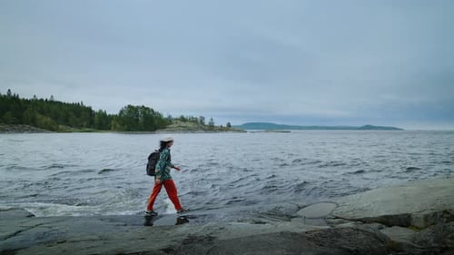 Travelling In Norway Tourist Woman With Backpack Walking Alone On Stone Shore Of Lake