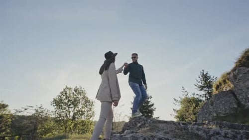 Stylish Woman and Man with Backpack Hiking on a Rocky Mountainside with Him Holding Her Hand to