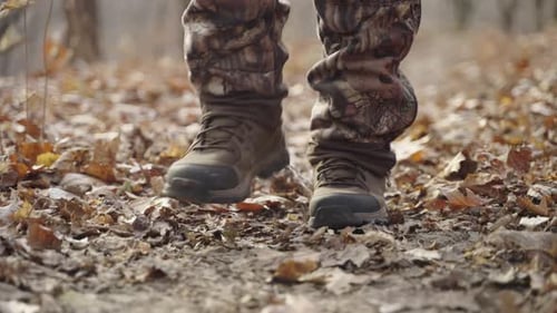 Hunter wearing boots and camouflage pants walking on winter forest path with dead leaves