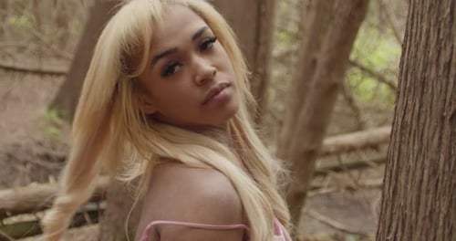 Black Girl With Long Blonde Hair Leaning Against Tree In Forest. She Flips Her Hair. Looks Up To Sky