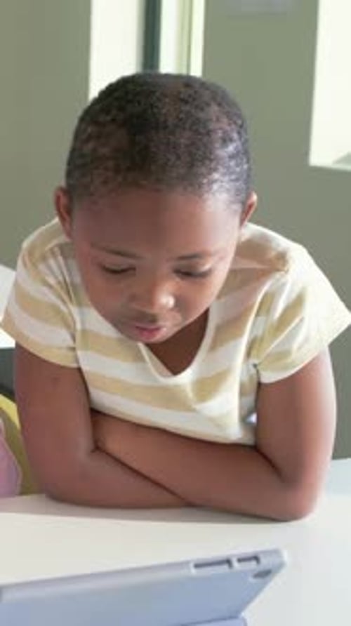 Vertical video: Leaning student in striped tee reading tablet content, tapping screen in classroom