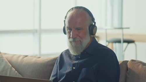 Senior Man Uses Laptop with Headphones on Couch