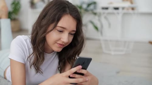 Junges fröhliches Mädchen, das drinnen auf dem Boden liegt und Smartphone benutzt