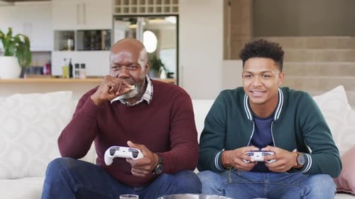 Excited african american father and adult son playing video game and celebrating, slow motion