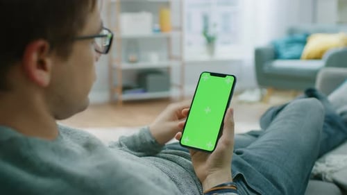 Man at Home Resting on a Couch using Smartphone with Green Mock-up Screen, Doing Swiping, Scrolling