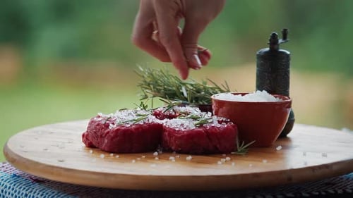 BBQ in the garden. Woman seasons filet mignon resting on the table with pepper and rosemary.