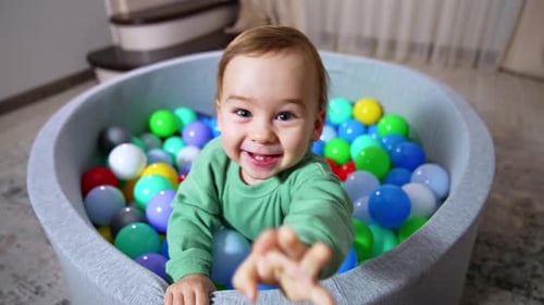 Joyful happy toddler smiling broadly stands in soft basin filled with colorful balls.
