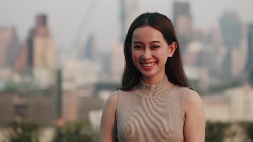 Portrait of Joyful Asian Woman in Casual Attire Smiles As Stands in Sunset City