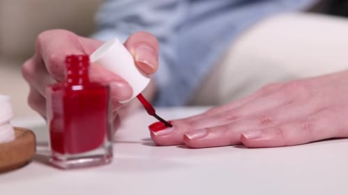 Woman applying red nail polish onto her nails at white table indoors, closeup