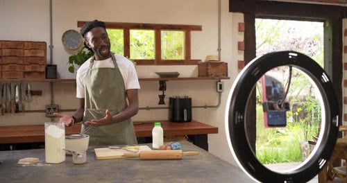 African American man in apron speaking to phone in ring light, recording recipe, measuring flour