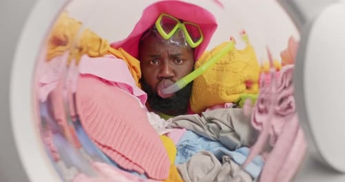 Un homme ethnique fait semblant de faire de la plongée avec tuba dans une machine à laver noyé dans du linge multicolore