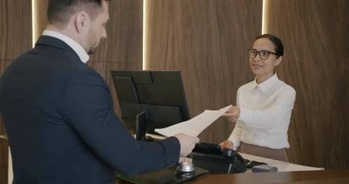 Young Businessman Talking to Receptionist Reading Documents in Hotel Lobby