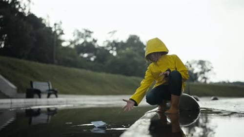 A Teenage Girl in a Yellow Jacket Floats a Paper Boat Through a Puddle Near a Curb in a Park