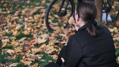 Young Woman Drinks Coffee on Lawn in Autumn Park Among Fallen Yellow Leaves