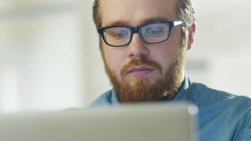 Portrait of a Bearded Young Man Wearing Glasses Sitting in His Office Working on a Computer. Comput