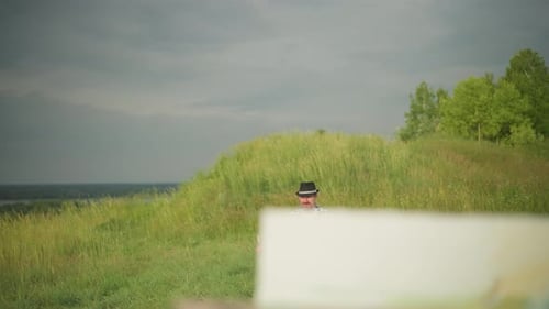 Man Holding Brush Seated in Sunlit Grassy Field Wearing Black Hat