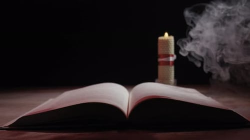 Offenes Buch auf einem Holztisch in der Nähe einer brennenden Kerze mit mystischem Rauch im Dunkeln
