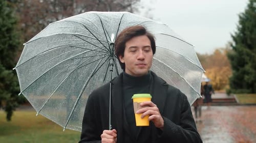 Young Man in a Coat Walking Under a Transparent Umbrella in the Autumn City Park Looking Around