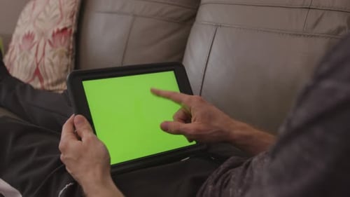 Male relaxing and sitting on the sofa tapping a tablet device with a green screen