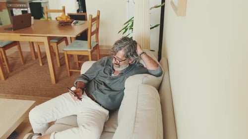 Elderly happy man using mobile phone while sitting on sofa at home. View from above