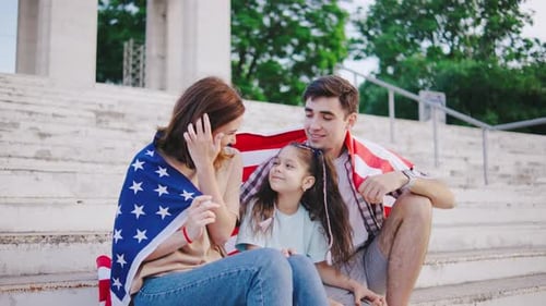 Portrait of A Young Family with a Child Girl is Sitting on the Steps and Wrapped in the Flag of the