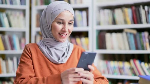 Close up. Muslim female student in hijab using smartphone in campus library. Happy girl looking