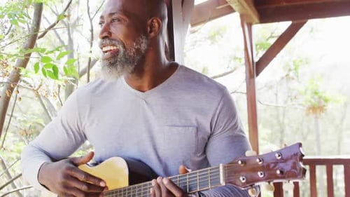 Happy senior african american man spending time in log cabin, playing guitar on balcony, slow motion