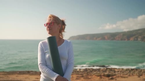 Pensioner Woman Holding Yoga Mat Standing on Ocean Shore