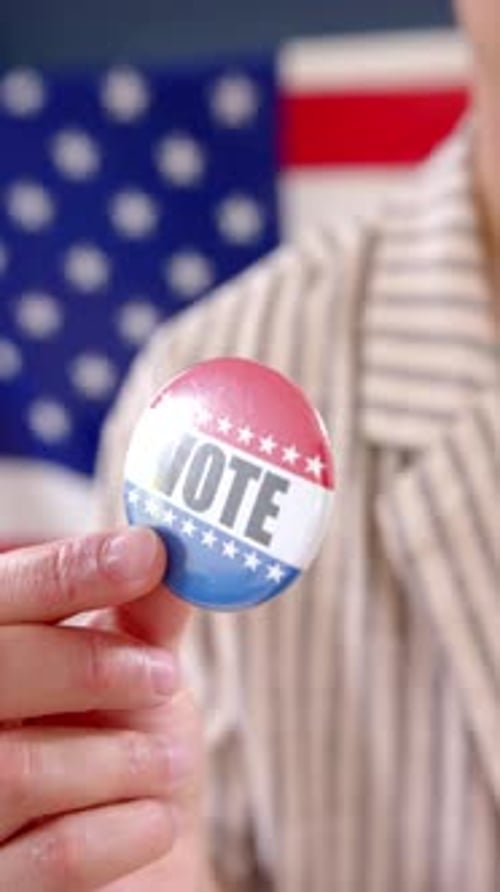 Hand Holds a VOTE Button in Front of Flag