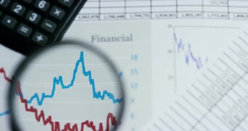 A hand with a magnifying glass passes over a financial newspaper and financial bag with a business