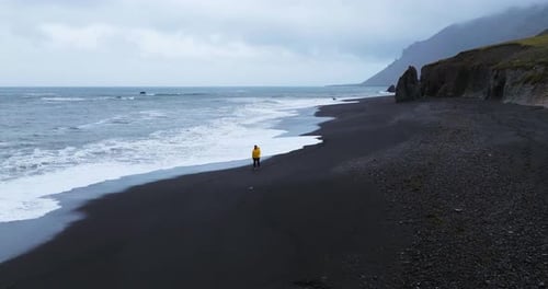 Man Walking In The Shoreline Of Lækjavik Black Sand Beach in Iceland. - aerial shot