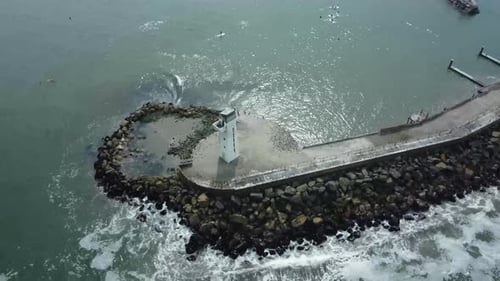 Drone orbiting around a white lighthouse at the end of a rocky pier. Waves splash on the rocks below