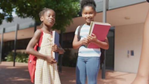 Video of three happy diverse schoolchildren with schoolbags greeting outside school