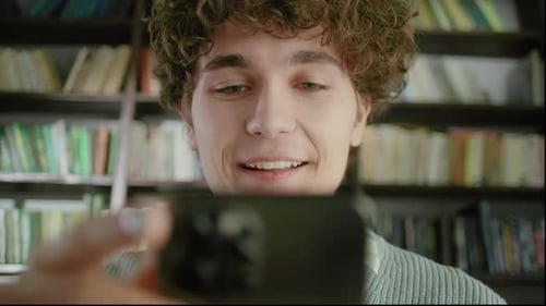 Portrait of Happy Young Student Chatting By Mobile Phone in University Library