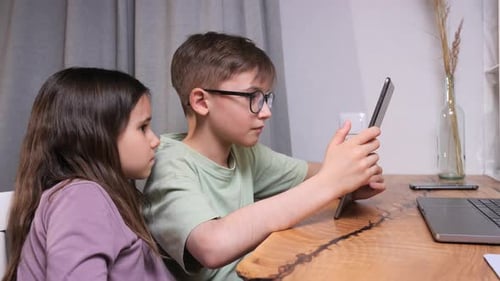 Two Children Boy and Girl Sitting at a Wooden Table Focused on a Tablet for Digital Learning and
