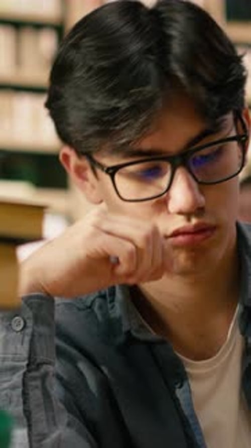 Vertical Video Exhausted Asian Student Leans on One Hand While Reading Open Book in Library